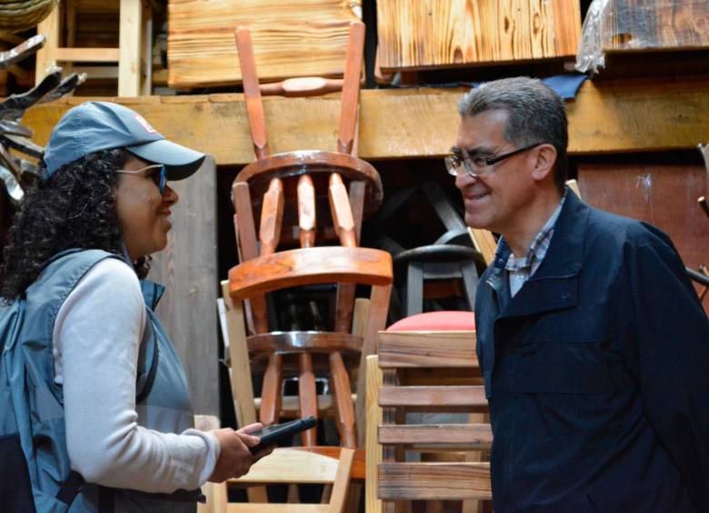 Una censista, con gorra y chaleco gris que sostiene una tableta electrónica, y un hombre conversan en un taller de carpintería con sillas de madera apiladas al fondo