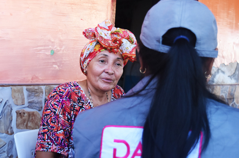 
                        Una mujer mayor con un pañuelo de colores en la cabeza conversando con una censista que se encuentra de espaldas que lleva una gorra y un chaleco con el logo del DANE